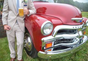 groom leaning up against a red truck holding a full beer glass